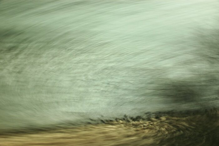 Bewegungsunscharfe Fotografie eines Flußufers, stark verwischte Flächen in blaugrau und braun am unteren Bildrand erkennbare Steine