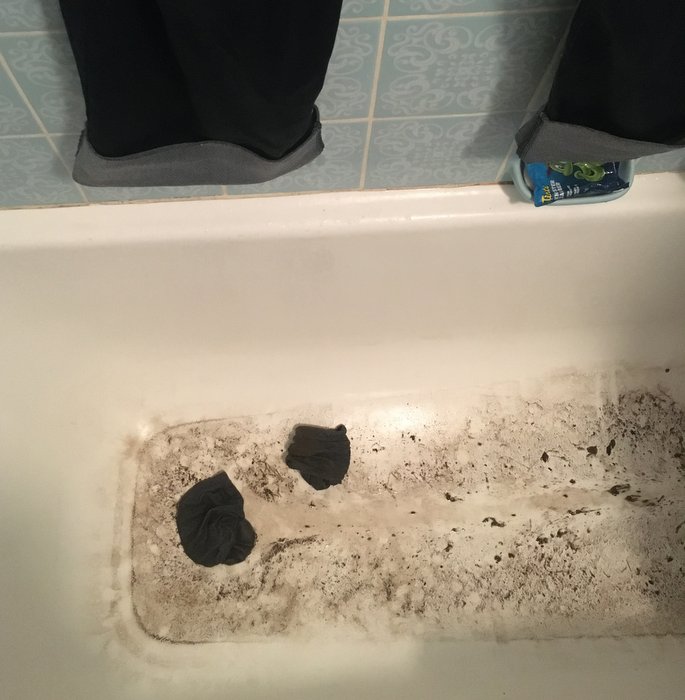 A completely filthy bathtub, you can see trousers hanging over it and crumpled socks lying in the tub.