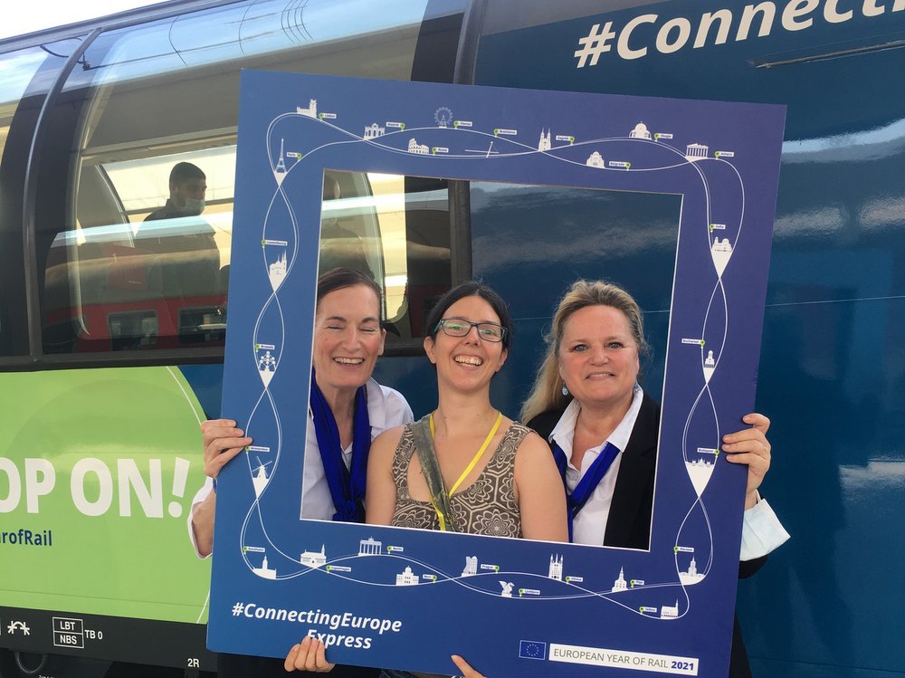 Drei Frauen blicken lachend durch einen Rahmen mit dem Design des Connecting Europe Express, der auch im Hintergrund zu sehen ist.