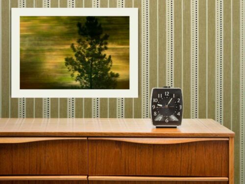 An old-fashioned bedside table with an analog alarm clock, on the wall behind it, there is a framed photograph of a coniferous tree in motion blur
