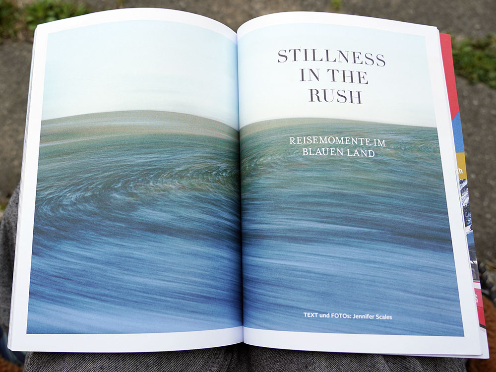 Eine aufgeschlagene Zeitschrift mit einem abstrakten Landschaftsbild über eine Doppelseite. Der Titel des Photoessays ist "Stillness in the Rush", Untertitel "Reisemomente im Blauen Land" "Fotos und Texte von Jennifer Scales"